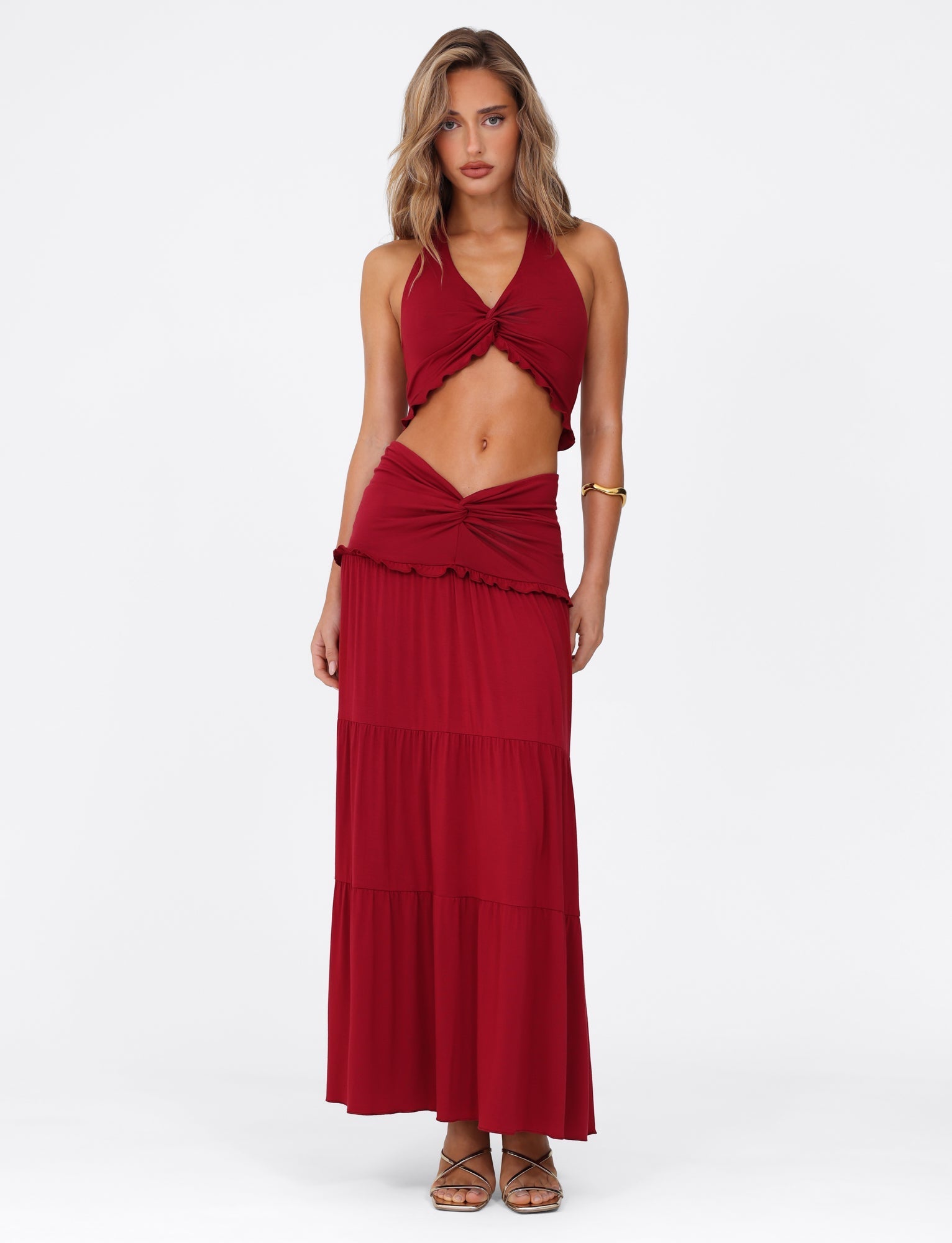 A woman poses in a fashionable two-piece ensemble, wearing the TIGERMIST KATYA MAXI SKIRT - RED and a matching red halter-top. Against a white background, she wears sandals and a gold bracelet. Her blonde hair flows in loose waves as she gazes directly at the camera.