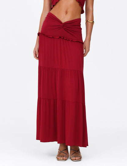 A person models the TIGERMIST KATYA MAXI SKIRT - RED, featuring a wide waistband and twist detail, paired with strappy sandals on a plain white backdrop.