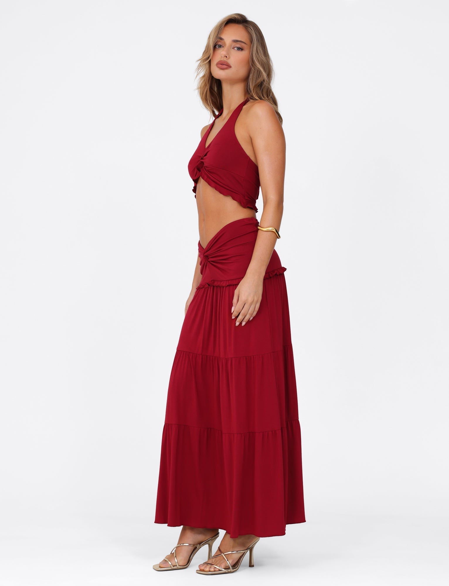 A woman models the TIGERMIST Katya Maxi Skirt - Red, paired with a red sleeveless crop top, on a white background. Her hair is down, and she accessorizes with strappy heels and a bracelet.