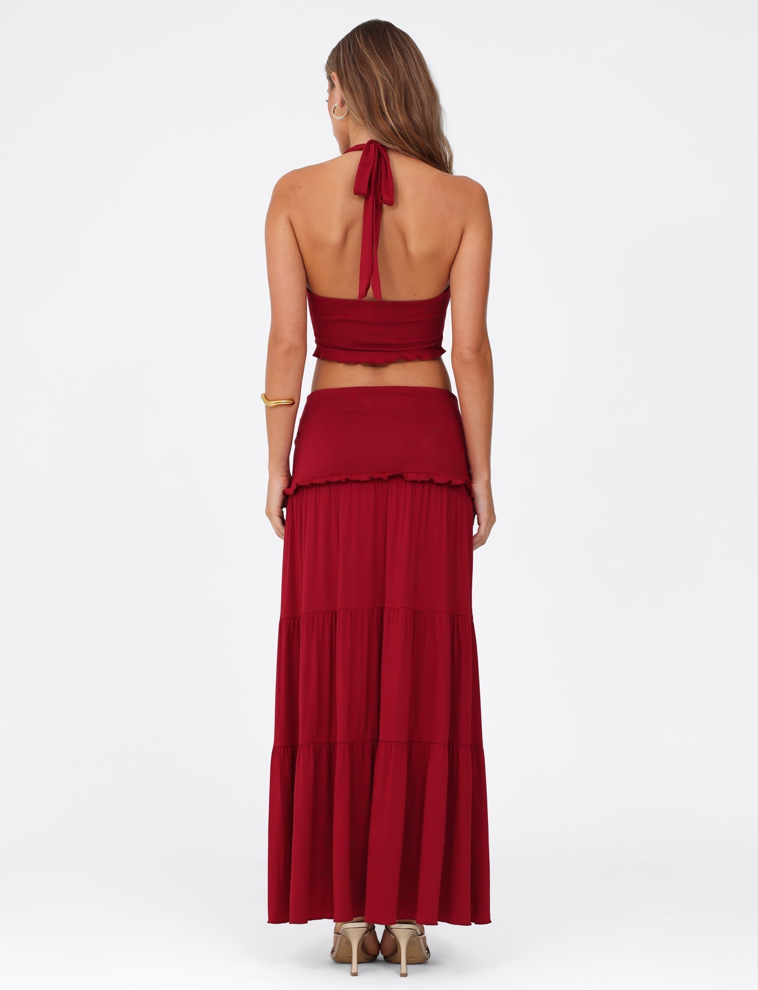 A woman with long hair stands facing away, wearing the TIGERMIST KATYA MAXI SKIRT - RED and a matching halter top. She accessorizes with a gold bracelet on her right wrist and beige heels. The plain white background accentuates her striking outfit and the skirt's tiered panels and wide waistband.